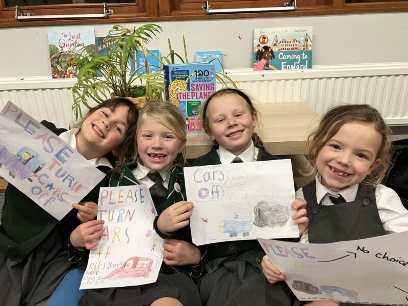 Four pupils holding posters encouraging parents to turn their car engines off when waiting